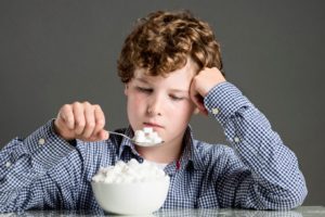 A BORED LOOKING CHILD LIFTS A SPOON OF SUGAR CUBES TO HIS MOUTH, FROM A HUGE BOWL OF SUGAR CUBES