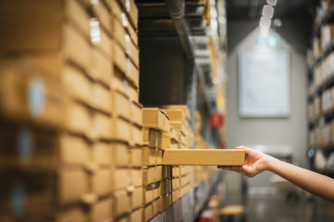 A HAND DRAWS A BOX OUT OF A STACK IN A WAREHOUSE RACK