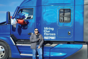 A WOMAN LEANS AGAINST A BLUE TRUCK, POINTING AT A DECAL ON ITS SIDE THAT READS HIRING OUR HEROES